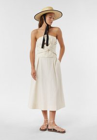 Beżowa sukienka bez ramiączek z przekręconym przodem, z pełną spódnicą, bocznymi kieszeniami, w połączeniu z płaskimi sandałami na paski i słomkowym kapeluszem.