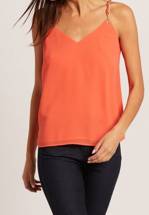 Vrouw in een mouwloze koraalkleurige top met dunne gouden schouderbandjes, gecombineerd met donkere, slim-fit jeans, staande tegen een effen achtergrond.