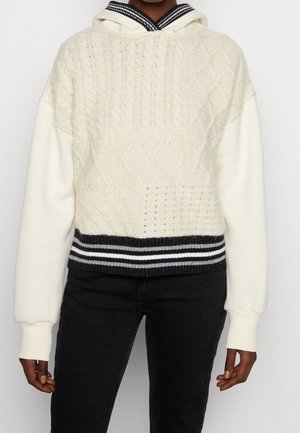 Personne portant un pull crème en maille torsadée avec un motif texturé, un ourlet côtelé à rayures noires et blanches contrastantes et des manches crème.