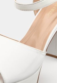Sandalia de tacón alto blanca con parte superior de cuero suave, diseño de punta abierta y una delgada correa en el tobillo. Presenta un tacón cuadrado y una plantilla en color beige.