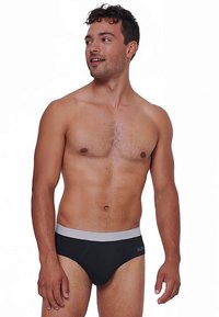 Uomo senza maglietta con capelli ricci corti, che indossa slip neri con una cintura grigia, che guarda leggermente a sinistra, in piedi contro uno sfondo bianco.