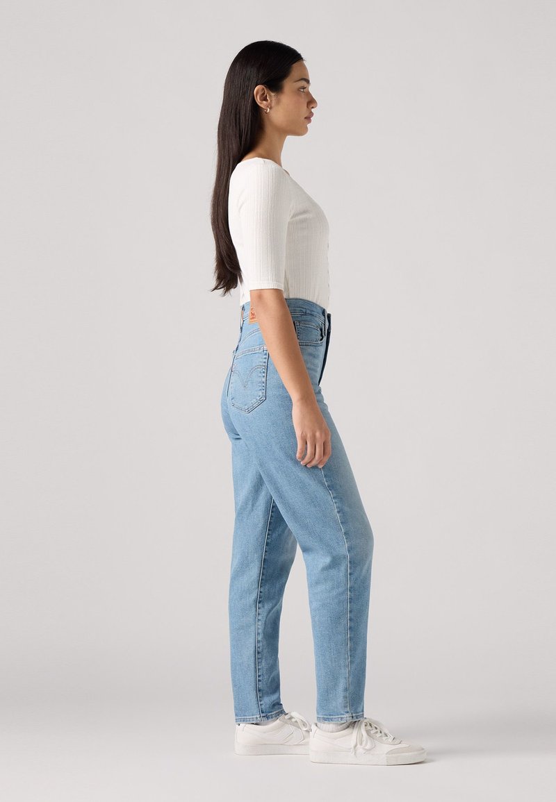 Hellblaue High-Waist-Jeans mit geradem Bein, kombiniert mit einem weißen, gerippten Langarmoberteil und weißen Sneakers.