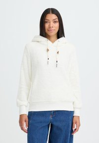 Balta, pūkuota džinsinė striukė su priekinėmis kišenėmis, kreminės spalvos juosta apačioje ir rankovėse, bei raišteliai su medinėmis ir spalvotomis detalėmis. Derinama su mėlynais džinsais.