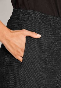 Hand rustend in de zak van zwarte broek met een subtiel wit stippenpatroon, gecombineerd met een donkere top.