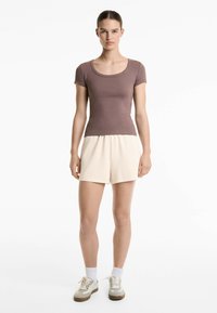 Top corto a righe marrone con collo rotondo, abbinato a pantaloni corti color crema. Il modello indossa calzini bianchi e sneakers beige.