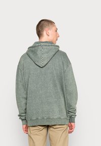 Felpa verde in cotone con una vestibilità rilassata, cappuccio con cordini e polsini a costine. Presenta una superficie morbida e testurizzata con una finitura sbiadita.