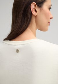 Helles Cremeshirt mit rundem Ausschnitt, strukturiertem Stoff und einem kleinen metallischen Logodetail hinten am Kragen.