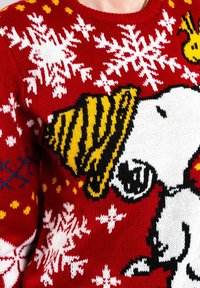 Roter Strickpullover mit weißen Schneeflocken und einem großen Snoopy-Design, der einen gelb gestreiften Partyhut trägt, mit schwarzen und gelben Akzenten.