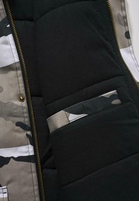 Chaleco acolchado negro tipo puffer con un forro interior de camuflaje, cremallera dorada y un pequeño bolsillo exterior con tejido de contraste.