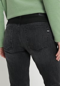Jeans di denim neri con una texture liscia, dotati di due tasche posteriori e un accento bianco sottile sulla tasca. Indossati con una maglietta in rete verde.