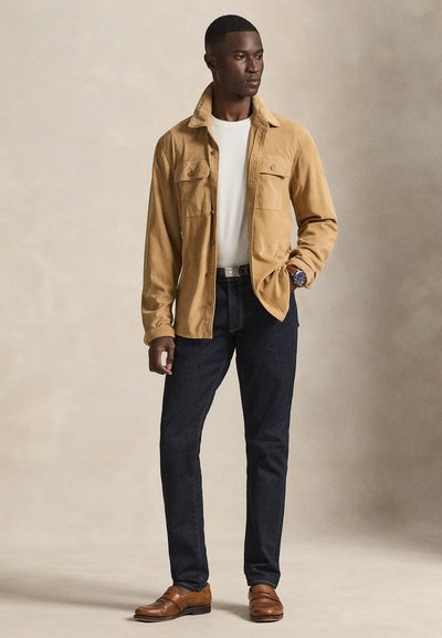 Veste-chemise en velours côtelé beige avec un design à boutons, t-shirt blanc en dessous, jean en denim foncé, et chaussures en cuir marron.