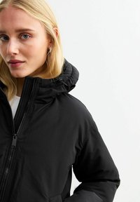 Chaqueta de plumas negra con capucha, cierre frontal, puños elásticos y una textura suave y brillante. Presenta una silueta ajustada.