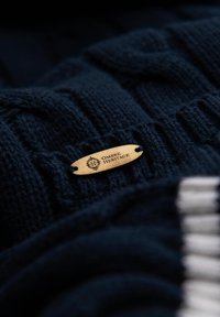 Maglione lavorato a maglia blu navy con un pattern testurizzato, con un'etichetta di legno che riporta la scritta "OMBRA HERITAGE" attaccata al tessuto.