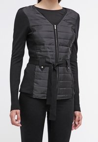 Kvinna i en svart vadderad zip-up jacka med långa ärmar och ett knytet tygbälte i midjan, tillsammans med svarta byxor.