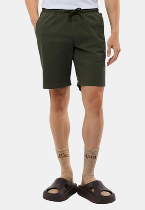 Shorts verts avec une taille élastique et un cordon de serrage, dotés de deux poches latérales. Associés à des chaussettes beige et des sandales noires à enfiler.