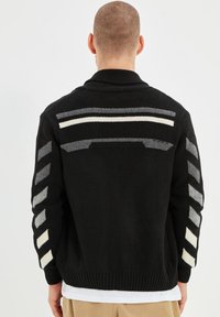 Cardigan nero lavorato a maglia con colletto, caratterizzato da righe orizzontali grigie e bianche sul retro e dettagli decorativi sulle maniche.
