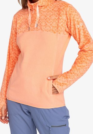 Vrouw draagt een perzikkleurige fleece trui met een patroon op het bovenstuk en de mouwen, duimgat-mouwen, en een blauwe cargobroek met een ritssluiting.