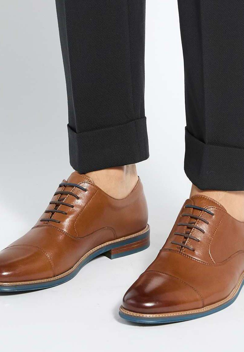 Chaussures de ville marron en cuir avec lacets foncés portées avec un pantalon noir retroussé, mettant en valeur des bouts de chaussures polis et des semelles superposées.