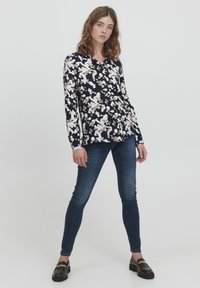Marinebluse med hvid og lys brun blomstermønster, lange ærmer, kravenedfald, parret med mørke denim jeans og sorte loafers.
