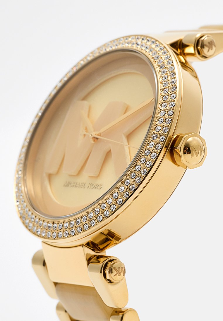 Montre dorée avec un boîtier rond, accents en cristal autour de la lunette, et logo "MK" en relief sur un fond clair. Liens de bracelet en métal.
