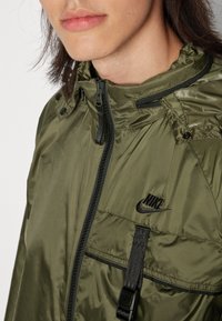 Olivgrön zip-up jacka med hög krage, justerbar huva och svart dragkedjeficka med Nike-logotyp. Slät, lättviktig tyg.