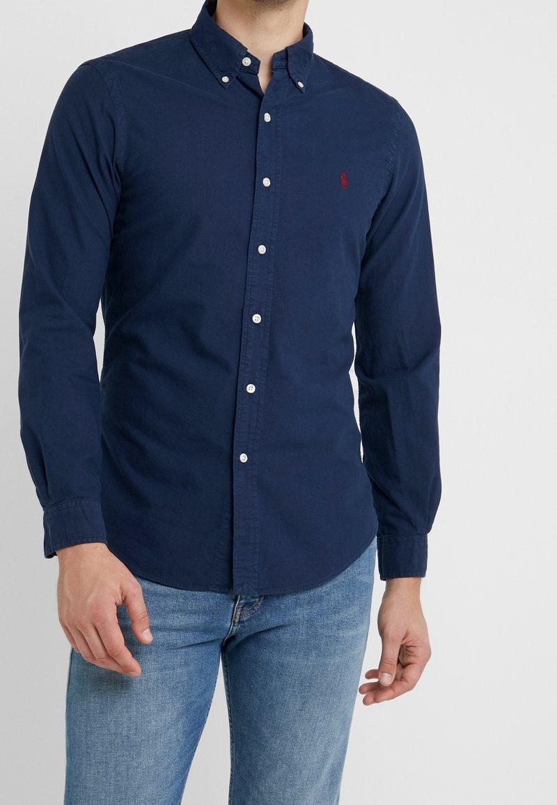 Námořnická modrá košile s dlouhými rukávy, klasickým límečkem a bílými knoflíky. Na levé hrudi je červené logo.