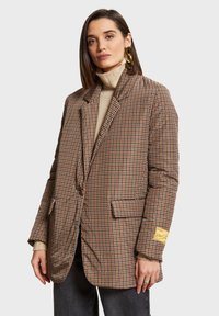 Tamni houndstooth blejzer s oversized krojem, s reverima, dva prednja džepa i oznakom branda na rukavu. Nosi se preko bež rolke.