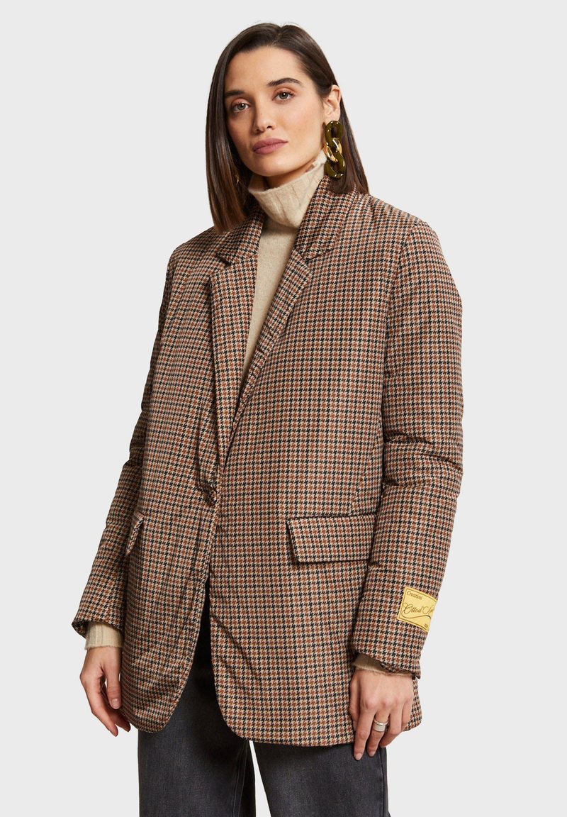 Tamni houndstooth blejzer s oversized krojem, s reverima, dva prednja džepa i oznakom branda na rukavu. Nosi se preko bež rolke.