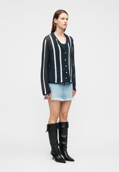 Γυναίκα που φοράει μαύρη και άσπρη ριγωτή ανοιχτή πλεκτή ζακέτα, ανοιχτό μπλε τζιν μίνι φούστα με ξεφτισμένα τελειώματα και μαύρες, ψηλές μποτάκια με τακούνι, στέκεται ακίνητη.