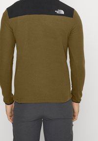 Chaqueta de forro polar verde oliva con panel negro en los hombros y logotipo. Mangas largas, escote redondo, textura suave y forma ajustada visible desde la vista trasera.