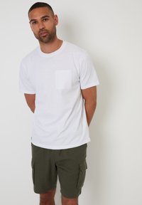 Ein weißes Baumwoll-T-Shirt mit kurzen Ärmeln und einer kleinen Brusttasche, kombiniert mit olivgrünen Cargo-Shorts mit Seitentaschen.