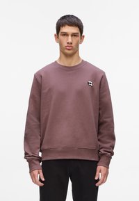 Sudadera marrón de tejido grueso, con cuello redondo y puños de canalé. Detalle notable: pequeño logo bordado en el pecho.