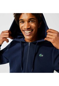 Sweat à capuche zippé bleu marine avec une capuche texturée, des cordons de serrage et un petit logo crocodile vert sur la poitrine. Tissu doux et coupe décontractée.