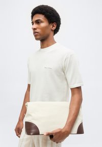 Άνδρας που φοράει κρεμ μπλουζάκι Marc O'Polo και ριγέ παντελόνια, κρατώντας μπεζ καμβά φάκελο με καφέ δερμάτινες γωνίες.