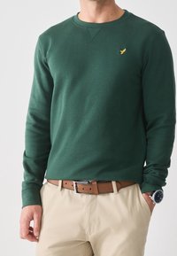 Felpa verde a maniche lunghe con scollo rotondo, decorata con un piccolo emblema dorato sul petto, abbinata a pantaloni beige e una cintura marrone.