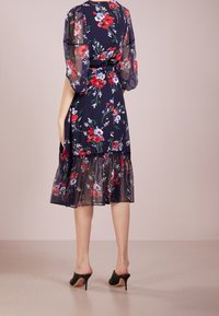 Mujer vestida con un vestido midi floral en azul marino con mangas abullonadas transparentes y dobladillo volado, combinado con zuecos de tacón negro, de pie sobre un suelo beige.