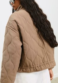 Femme aux longs cheveux ondulés portant une grande veste matelassée beige et un pantalon blanc, vue de dos sur fond uni.
