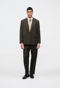 Hnedé dvojradové sako, zodpovedajúce nohavice, krémová pletená vesta, biela košeľa, hnedá kravata. Lesklé hnedé topánky. Hladká látka, strihané na mieru.