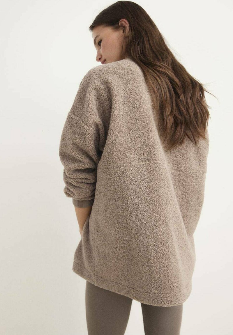 Vrouw met lang bruin haar, gekleed in een losse, beige fleecjas en taupekleurige leggings, staande opzij tegen een plain witte achtergrond.