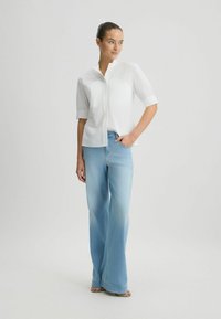 Chemise blanche à manches courtes et col montant, associée à un jean ample bleu clair, montrant un tissu lisse et léger.
