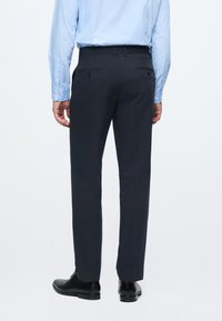 Dunkelblaue Hosen mit glatter Textur, ausgestattet mit zwei Hintertaschen mit Knöpfen. Schwarze elegante Schuhe runden das Outfit ab.