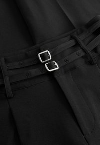 Két keskeny fekete szövetöv ezüst négyszögletes csattal, fekete pliszírozott anyagra rögzítve.