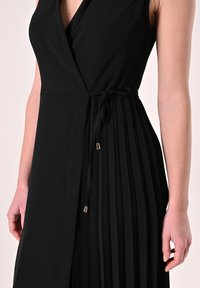 Zwarte mouwloze jurk met een wikkelontwerp, voorzien van plooien aan één zijde, een ceintuur met strik, en goudkleurige sierdelen aan de uiteinden.