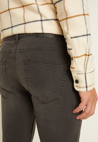 Jeans de ganga cinza com uma textura suave, apresentando dois bolsos traseiros e uma etiqueta preta na cinta, usados com uma camisa de flanela xadrez.