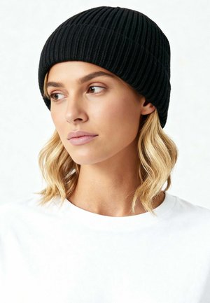 Donna con capelli biondi ondulati, che indossa un berretto nero a coste e una maglietta bianca, che guarda leggermente verso sinistra su uno sfondo semplice.