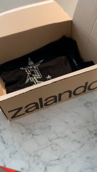 Καφέ χαρτόκουτο με λογότυπο Zalando, που περιέχει ένα μαύρο πουκάμισο με σχέδιο αστεριού από ασημί κρυστάλλους και ορατή υφή υφάσματος.