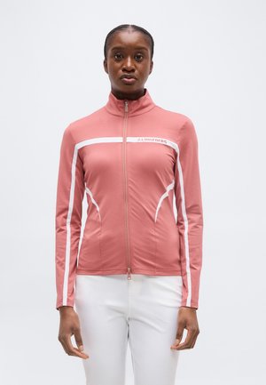 Femeie purtând o jachetă roz prăfuit cu fermoar, cu dungi albe și pantaloni albi, stând pe un fundal simplu, deschis la culoare.