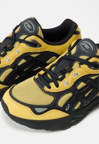 Τα αθλητικά παπούτσια Asics διαθέτουν σχεδιασμό με κίτρινο και μαύρο δίχτυ, μαύρα κορδόνια, λαστιχένια σόλα και μεταλλικές λεπτομέρειες στο πάνω μέρος.