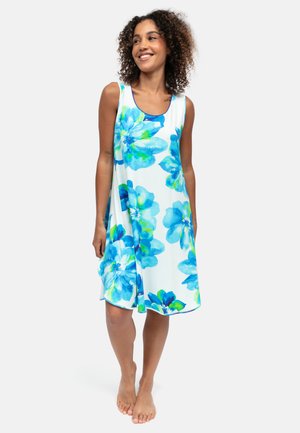 Kobieta z kręconymi włosami, uśmiechnięta, ubrana w białą, bezrękawnikową sukienkę z dużymi niebiesko-zielonymi kwiatowymi wzorami, stojąca boso na jednolitym tle.