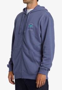 Sudadera con capucha y cremallera en suave tela azul. Cuenta con un bolsillo tipo canguro en el frente y un logo gráfico con diseño de palmera en el pecho.